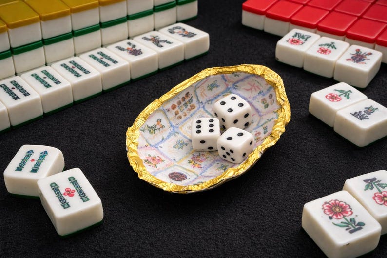 May include: An oval-shaped decorative dish with a gold-toned, textured rim and a white interior featuring a grid of small, colorful floral and patterned illustrations. Three white six-sided dice with black pips rest inside the dish.