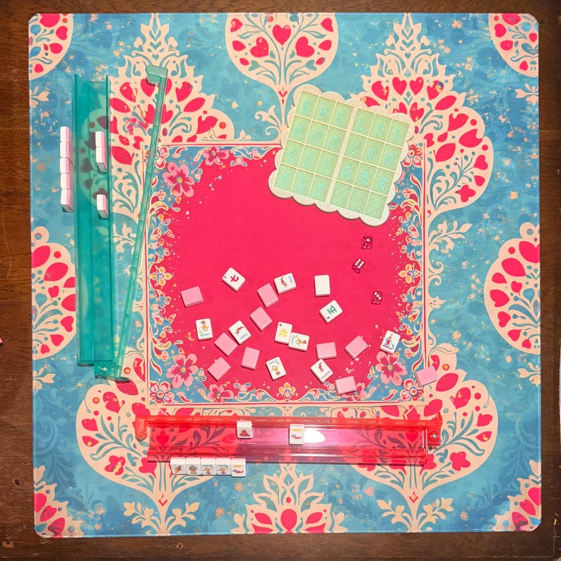 May include: A Mahjong game set on a patterned mat. The set includes pink tiles, dice, and racks. The mat has a pink center and a blue floral design. Green and red plastic pieces are also visible.