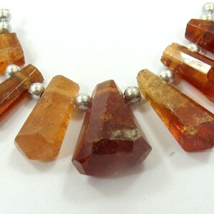 May include: A collection of faceted, amber-colored gemstone beads with silver-colored metal accents. The gemstones vary in shape, with some being elongated and others more geometric. The beads are strung together, suggesting a necklace or bracelet.