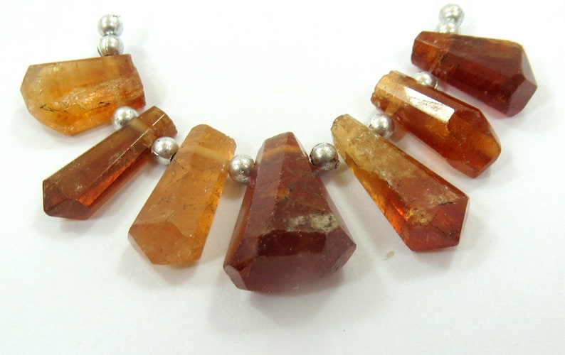 May include: A collection of faceted, amber-colored gemstone beads with silver-colored metal accents. The gemstones vary in shape, with some being elongated and others more geometric. The beads are strung together, suggesting a necklace or bracelet.