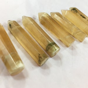 May include: Seven golden-yellow crystal wands are arranged diagonally. Each wand has a faceted, rectangular shape with a pointed tip. Some wands have green and black inclusions. The crystals are displayed on a white surface.