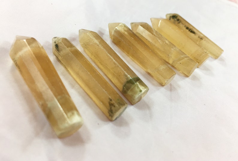 May include: Seven golden-yellow crystal wands are arranged diagonally. Each wand has a faceted, rectangular shape with a pointed tip. Some wands have green and black inclusions. The crystals are displayed on a white surface.
