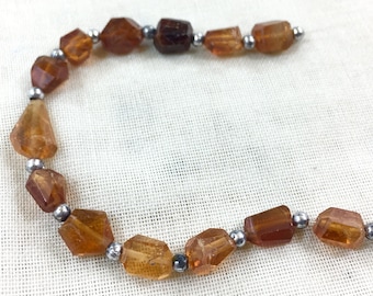May include: A necklace featuring faceted, amber-colored gemstones strung together with small, silver-colored beads. The gemstones have a variety of shapes and sizes, and the necklace is displayed on a white, textured surface.