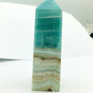 May include: A rectangular crystal obelisk with a pointed top. The crystal displays bands of turquoise, white, and tan colours. The polished surface reflects light, highlighting the natural patterns and textures of the stone.
