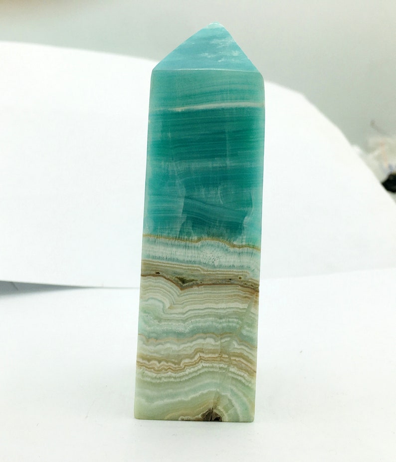 May include: A rectangular crystal obelisk with a pointed top. The crystal displays bands of turquoise, white, and tan colours. The polished surface reflects light, highlighting the natural patterns and textures of the stone.