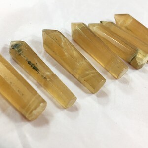 May include: Seven golden-yellow crystal wands with pointed tips are arranged diagonally on a white surface. The crystals have a translucent appearance with subtle variations in color and some dark inclusions.
