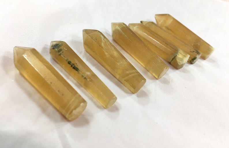 May include: Seven golden-yellow crystal wands with pointed tips are arranged diagonally on a white surface. The crystals have a translucent appearance with subtle variations in color and some dark inclusions.