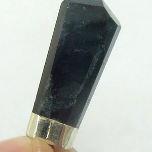May include: A dark, faceted crystal pendant with a silver-colored metal band at the base. The crystal is a deep, opaque black with subtle variations in color. The pendant is held between fingers against a white background.
