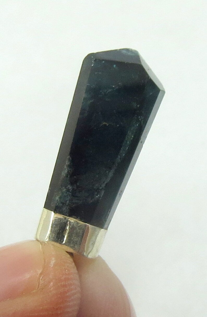 May include: A dark, faceted crystal pendant with a silver-colored metal band at the base. The crystal is a deep, opaque black with subtle variations in color. The pendant is held between fingers against a white background.