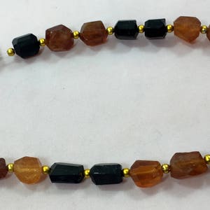 May include: A necklace featuring alternating black and amber-colored gemstone beads, interspersed with small gold-colored beads. The gemstones have a faceted, irregular shape. The necklace is strung on a thin, clear cord.
