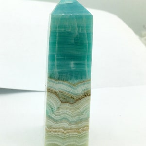May include: A rectangular, polished crystal obelisk. The top is a pointed pyramid shape. The crystal has layers of teal, white, and tan colours. The obelisk is standing upright against a white background.