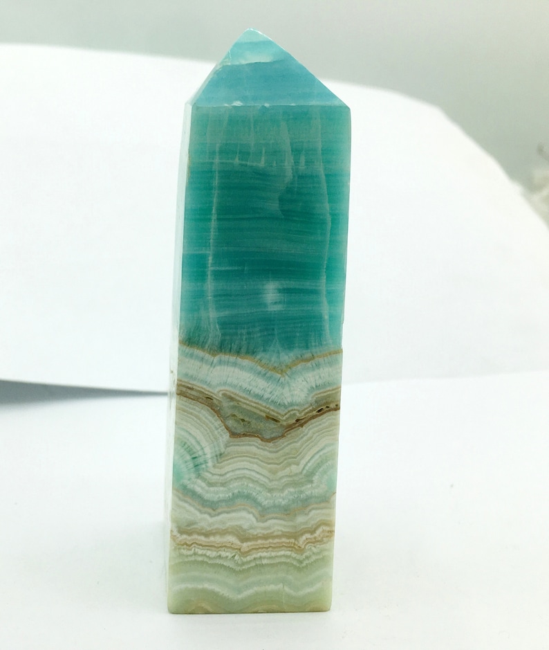 May include: A rectangular, polished crystal obelisk. The top is a pointed pyramid shape. The crystal has layers of teal, white, and tan colours. The obelisk is standing upright against a white background.