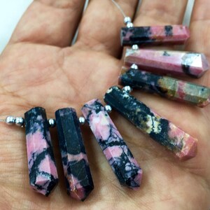 May include: Close-up of several polished, hexagonal-shaped gemstones. The stones feature a mix of pink, black, and white colors. Each stone is strung on a thin, silver-colored wire with small, silver beads.