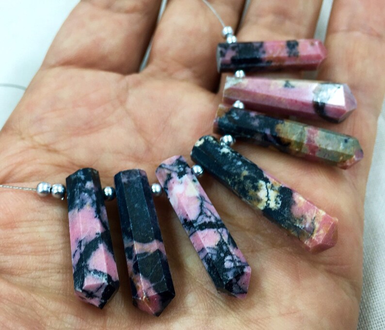May include: Close-up of several polished, hexagonal-shaped gemstones. The stones feature a mix of pink, black, and white colors. Each stone is strung on a thin, silver-colored wire with small, silver beads.