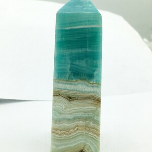 May include: A polished, rectangular crystal tower with a flat top. The crystal features bands of teal, light blue, and tan colours. The tower is standing upright against a white background.