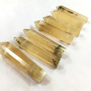 May include: Six golden-yellow crystal wands with pointed tips are arranged diagonally on a white surface. The crystals have a translucent quality, with some showing subtle internal patterns and dark green inclusions near the base. These are likely used for spiritual practices.
