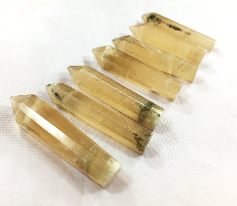 May include: Six golden-yellow crystal wands with pointed tips are arranged diagonally on a white surface. The crystals have a translucent quality, with some showing subtle internal patterns and dark green inclusions near the base. These are likely used for spiritual practices.