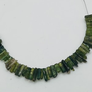 May include: A necklace of green tourmaline beads. The beads are cylindrical and vary in shades of green, from dark to light. The beads are strung on a thin, silver-colored wire.