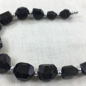 Puede incluir: Un collar de cuentas de piedras preciosas negras facetadas, ensartadas con pequeñas cuentas plateadas. Las piedras preciosas tienen una forma geométrica y una superficie pulida. Las cuentas se utilizan probablemente para la fabricación de joyas.