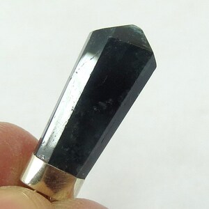 May include: A black, faceted crystal with a silver-colored metal base. The crystal is a dark, opaque color and has a polished, reflective surface. The crystal is held by a person's fingers.