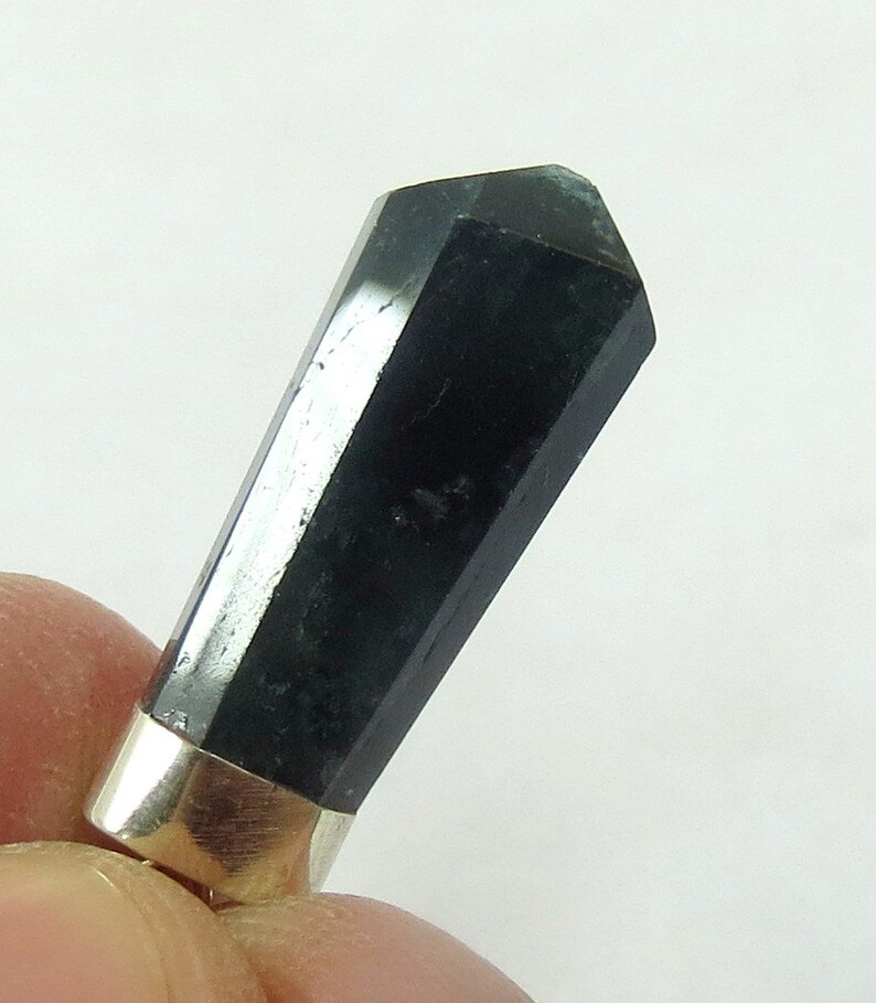 May include: A black, faceted crystal with a silver-colored metal base. The crystal is a dark, opaque color and has a polished, reflective surface. The crystal is held by a person's fingers.