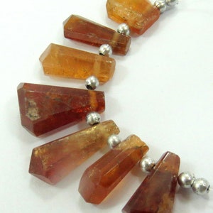May include: A strand of faceted, amber-colored gemstone beads with silver-colored spacer beads. The gemstones have a natural, crystalline appearance and vary in size and shape. The beads are strung on a clear, thin cord, suitable for jewelry making.