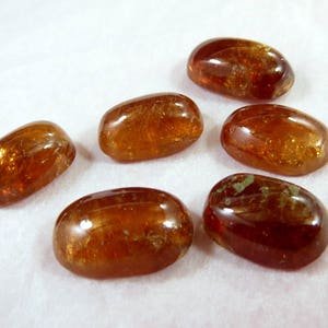 Pode incluir: Seis pedras preciosas ovais polidas, em tons âmbar e castanho, dispostas sobre uma superfície branca. As pedras têm uma textura lisa e brilhante, algumas exibindo padrões internos e inclusões. As pedras preciosas variam ligeiramente em cor e tamanho.