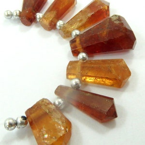 May include: A necklace featuring several faceted, amber-colored gemstone beads. The beads are strung together with small, silver-colored metal beads. The gemstones have a variety of shapes, with some appearing more rectangular and others more irregular.