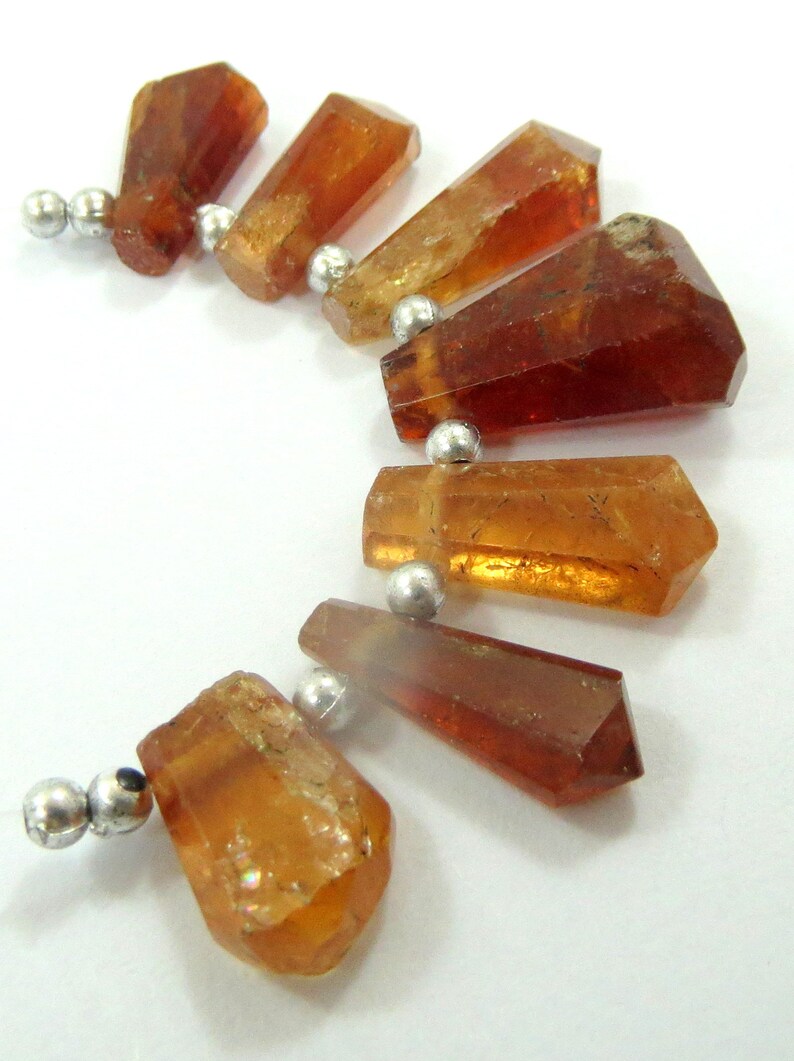 May include: A necklace featuring several faceted, amber-colored gemstone beads. The beads are strung together with small, silver-colored metal beads. The gemstones have a variety of shapes, with some appearing more rectangular and others more irregular.