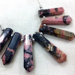 May include: A collection of faceted, elongated gemstone beads in shades of pink, black, and brown, strung on a thin silver wire. The beads have a polished, hexagonal shape and are arranged in a necklace-like pattern.