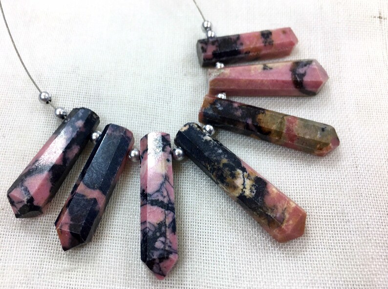 May include: A collection of faceted, elongated gemstone beads in shades of pink, black, and brown, strung on a thin silver wire. The beads have a polished, hexagonal shape and are arranged in a necklace-like pattern.