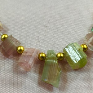 May include: A necklace featuring a series of rough-cut tourmaline gemstones in shades of pink, green, and clear, interspersed with small gold-colored beads. The gemstones are strung on a thin, white cord, creating a delicate and natural aesthetic.