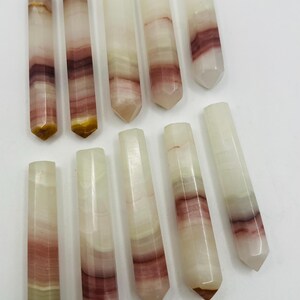 May include: Ten polished onyx crystal wands are displayed. The wands are rectangular with a pointed end. The colours are white, cream, burgundy, and brown. The wands are approximately 10 cm long.