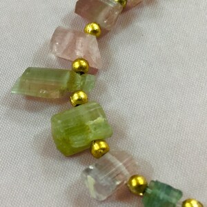 May include: A necklace featuring a strand of rough-cut, multi-colored tourmaline gemstones. The stones are in shades of pink, green, and clear, separated by small gold-colored beads. The necklace is strung on a thin, clear cord.
