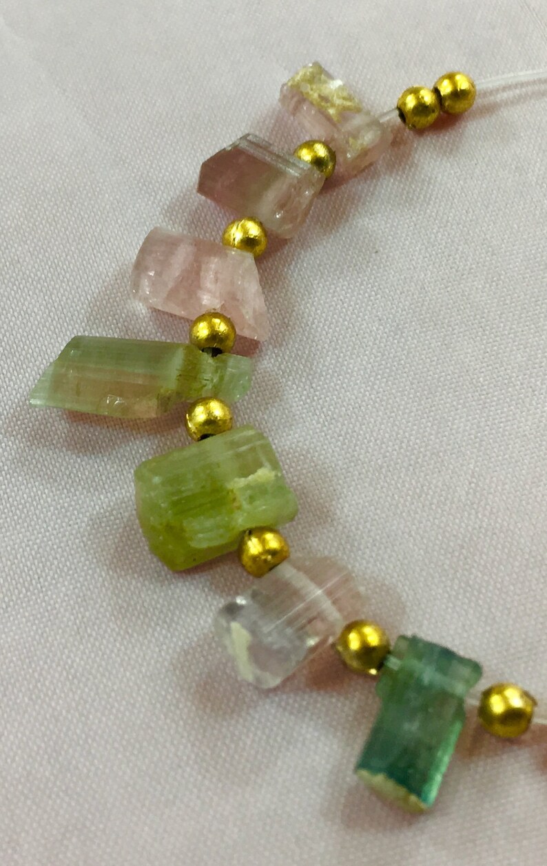 May include: A necklace featuring a strand of rough-cut, multi-colored tourmaline gemstones. The stones are in shades of pink, green, and clear, separated by small gold-colored beads. The necklace is strung on a thin, clear cord.