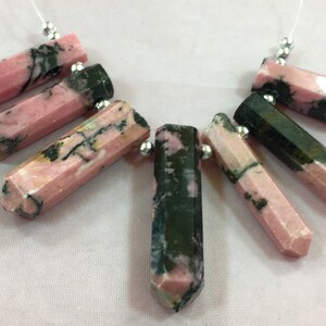 May include: A collection of faceted, elongated gemstone pendants. The stones are primarily pink with dark green and black veining. Each pendant is attached to a silver bead and a thin white string, suggesting they are for jewelry making.