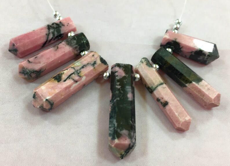 May include: A collection of faceted, elongated gemstone pendants. The stones are primarily pink with dark green and black veining. Each pendant is attached to a silver bead and a thin white string, suggesting they are for jewelry making.