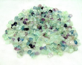 May include: A pile of tumbled fluorite stones in shades of green, purple, blue, and clear. The stones are small and irregular in shape, with a polished, glossy appearance. The stones are scattered on a white surface.
