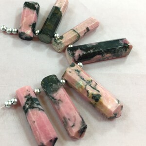 May include: Pink and black gemstone beads with a faceted, hexagonal shape. The beads are strung on a white string with silver-colored beads. The gemstones have a mix of pink and dark green colors.