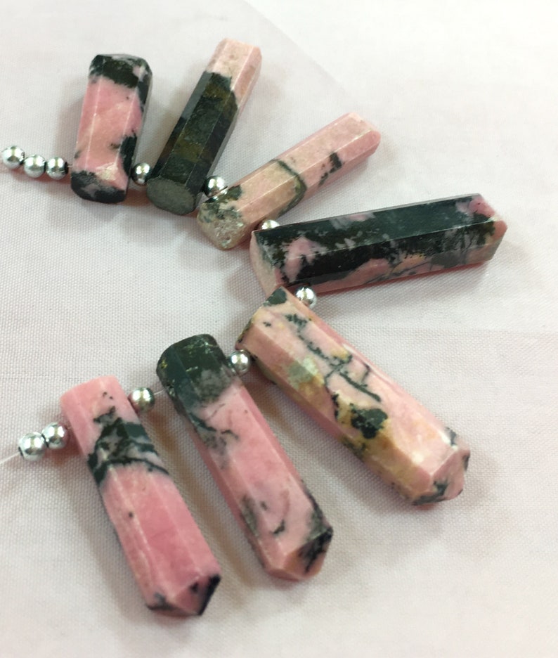 May include: Pink and black gemstone beads with a faceted, hexagonal shape. The beads are strung on a white string with silver-colored beads. The gemstones have a mix of pink and dark green colors.
