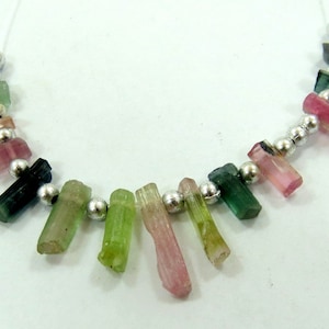 May include: A necklace featuring a strand of colorful tourmaline crystals in shades of green, pink, and black. The crystals are separated by small, silver-colored beads. The necklace is strung on a white cord.