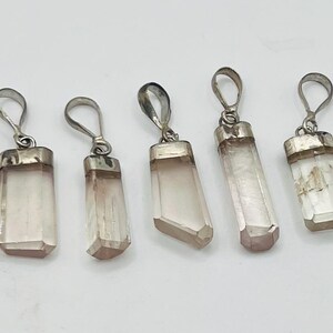 May include: Five crystal pendants with silver-colored metal loops and caps. The crystals are a pale pink and white color, with a rectangular or faceted shape. The pendants are displayed against a white background.