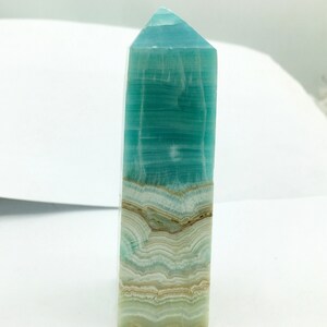 May include: A polished, rectangular crystal obelisk with a pointed top. The crystal features layers of teal, white, and tan colours, creating a striped pattern. The obelisk is standing upright against a white background.
