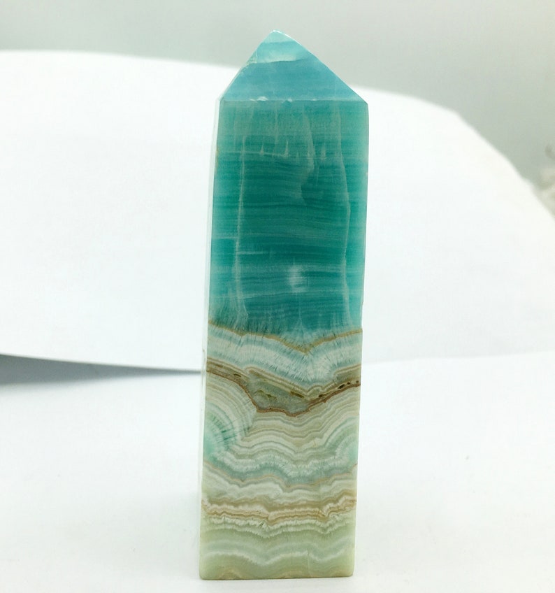 May include: A polished, rectangular crystal obelisk with a pointed top. The crystal features layers of teal, white, and tan colours, creating a striped pattern. The obelisk is standing upright against a white background.