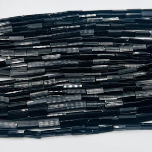 May include: Close-up of a collection of black tourmaline beads. The beads are rectangular and have a polished, glossy finish. The beads are strung together, creating a dense, textured surface. The overall color is a deep, solid black, with subtle variations in tone.