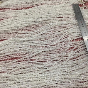 May include: Strands of clear, faceted beads, possibly quartz or crystal, are arranged in wavy lines. A metal ruler with centimeter markings is placed diagonally across the beads, indicating a measurement of approximately 30 cm. The beads are strung together with silver-colored wire.