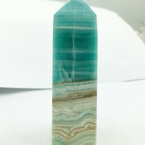May include: A rectangular crystal obelisk with a pointed top. The obelisk displays layers of turquoise, white, and tan hues. The crystal has a polished surface and a natural, layered appearance, suitable for home decor or spiritual practices.