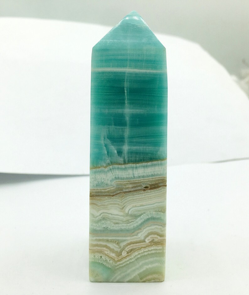May include: A rectangular crystal obelisk with a pointed top. The obelisk displays layers of turquoise, white, and tan hues. The crystal has a polished surface and a natural, layered appearance, suitable for home decor or spiritual practices.