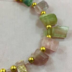 May include: A necklace featuring a strand of multi-colored tourmaline gemstones in shades of green, pink, and purple, separated by small gold beads. The gemstones are irregularly shaped and have a natural, raw appearance.