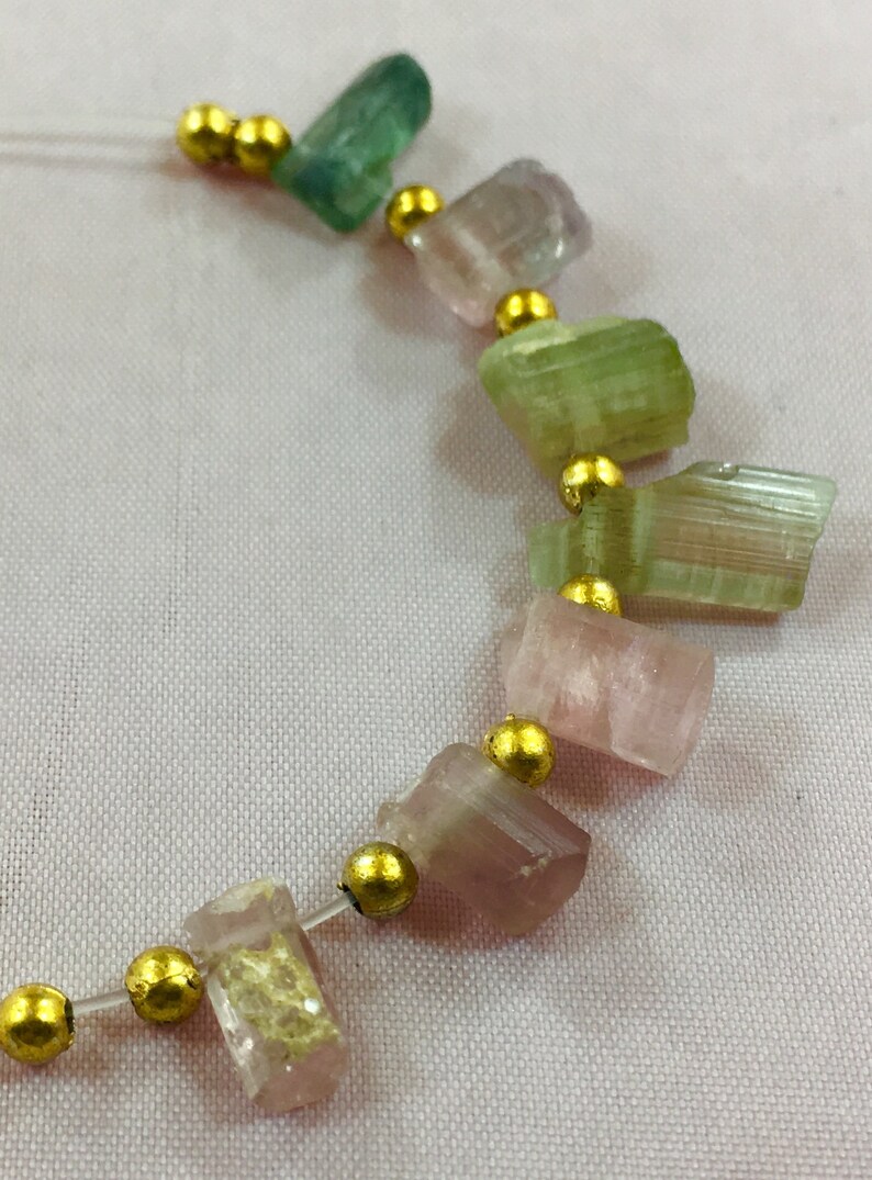 May include: A necklace featuring a strand of multi-colored tourmaline gemstones in shades of green, pink, and purple, separated by small gold beads. The gemstones are irregularly shaped and have a natural, raw appearance.